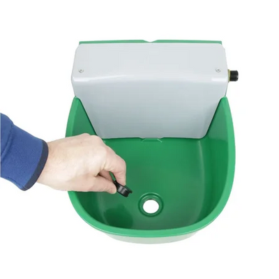 Nahaufnahme einer Hand mit blauem Ärmel, die einen kleinen schwarzen Knopf in einem grünen Kunststoffbecken betätigt; in der Mitte ist eine Abflussöffnung sichtbar, darüber sitzt ein weisser Kasten.