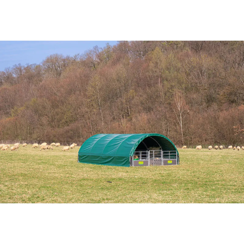 Grüner, gewölbter Viehstall aus Plane auf einer grasbewachsenen Weide; im Hintergrund eine Schafherde und ein bewaldeter Hang.
