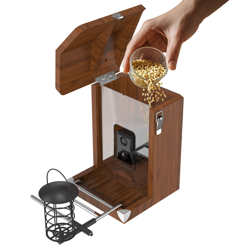 Holzbox mit geöffnetem Deckel; eine Hand giesst Haferflocken aus einem Glasbehälter in den oberen Innenraum, sichtbar durch eine transparente Seitenwand; ein Metallkorb ist am unteren Rand des Geräts sichtbar.