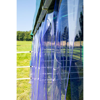 Seitenansicht einer langen Wand aus blauen transparenten Kunststofffolien an einem grünen Rahmen, mit Grasland, weissem Zaun und Baumreihe im Hintergrund.