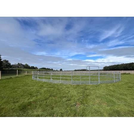 Ovale Koppel aus Metallzaunpaneelen auf einer grünen Weide; offene Landschaft mit Bäumen und blauem Himmel im Hintergrund.