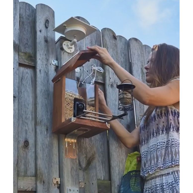 Frau montiert an einem Holzzaun eine selbstgebaute Vogelhaus-Futterstation; die Box hat ein Dach, eine transparente Front und eine integrierte Kamera, daneben hängt eine Laterne.