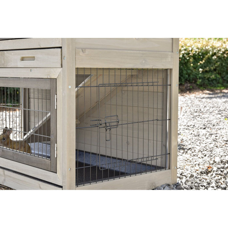 Holz-Hundehütte mit Drahtgitterfront im Aussenbereich; ein kleiner Hund blickt aus dem Käfig, neben Kiesweg.