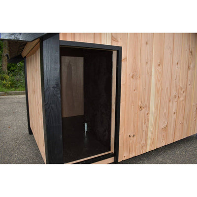 Holz-Hundehütte mit offener Tür, dunklem Innenraum, Aussenwand aus Naturholzplatten und schwarzem Türrahmen, steht auf einer Asphaltfläche.