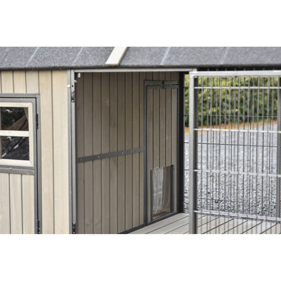 Holzschuppen mit vertikalen Brettern, links kleines Fenster, mittig Tür, rechts Metallgitterzaun, Kiesboden davor.