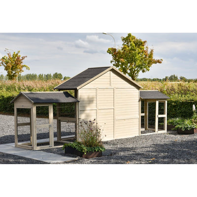 Grosser Holz-Hühnerstall mit schwarzen Dächern, seitliche Anbauten und Drahtgehege, steht auf Kiesboden im Garten; Hintergrund mit Bäumen und Feldern.
