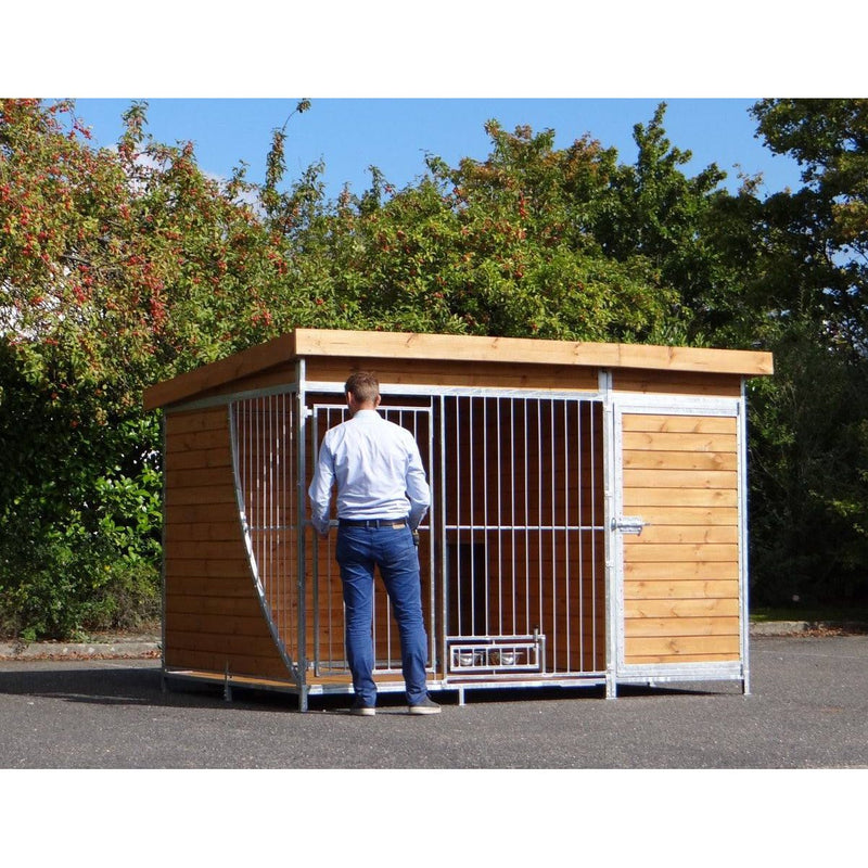 Mann in Jeans und hellblauem Hemd steht vor einem grossen Hundezwinger aus Holz und Metallstäben im Freien.