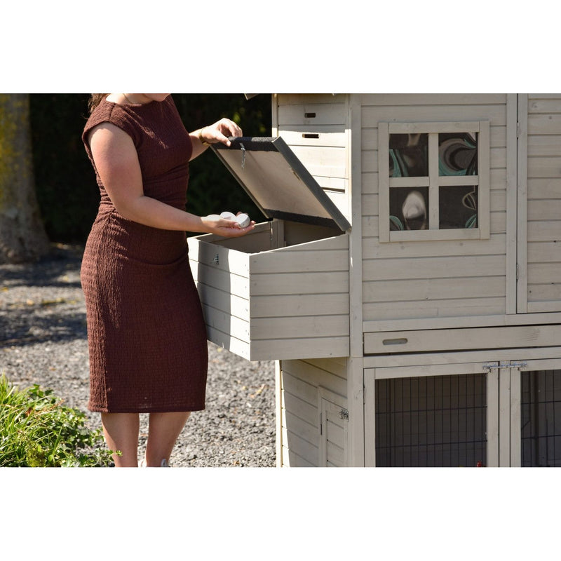Frau in braunem Kleid holt frische Eier aus dem geöffneten Nistkasten eines weissen Holzhühnerstalls im Freien.