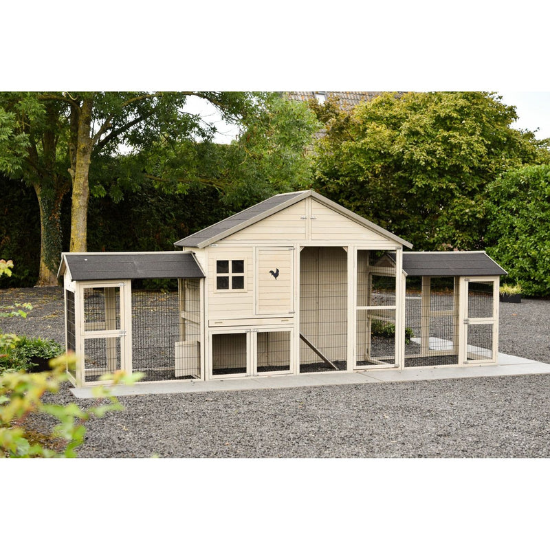 Grosser Hühnerstall aus Holz mit mehreren Abteilen und Drahtgehege, zentralem Gebäudeteil und seitlichen Ausläufen, auf Kiesboden in einem Garten mit Bäumen.