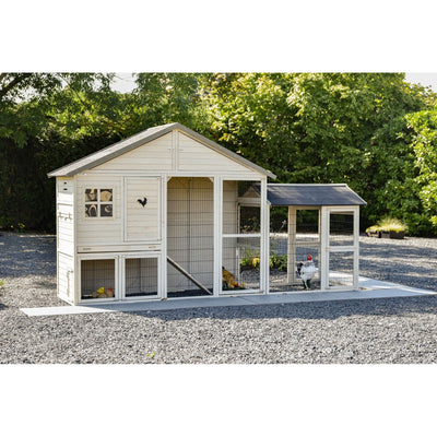 Weisses Holzhühnerhaus mit Drahtgehege und separatem Auslauf aus Drahtnetz, auf Kiesboden, umgeben von grünen Bäumen.