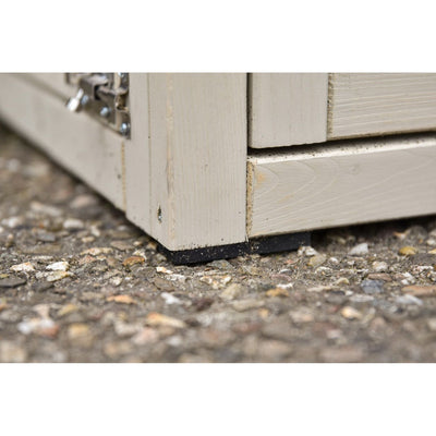 Nahaufnahme der unteren Ecke eines hellbeigen Holz-Gartenhauses/Schuppens mit Metall-Scharnier am linken Rand; der Holzfuss ruht auf einem schwarzen Unterlegstück auf Kiesboden.