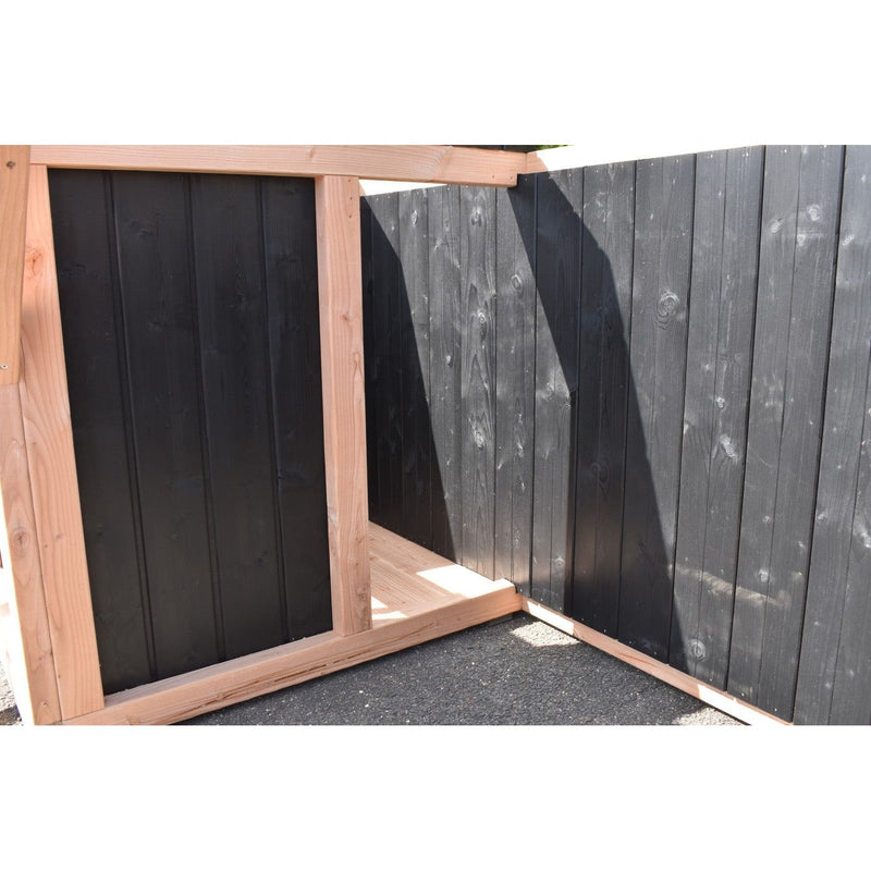 Eckzaun aus dunklen Holzbrettern mit hellem Holzrahmen; Gartenbereich, Asphaltboden und Schattenwurf der Zaunstruktur.