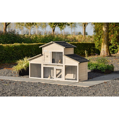 Mehrstöckiger Kaninchenstall aus hellem Holz mit Drahtgittertüren und schwarzem Schindeldach, im Garten auf Kiesboden, umgeben von Hecken und Bäumen.