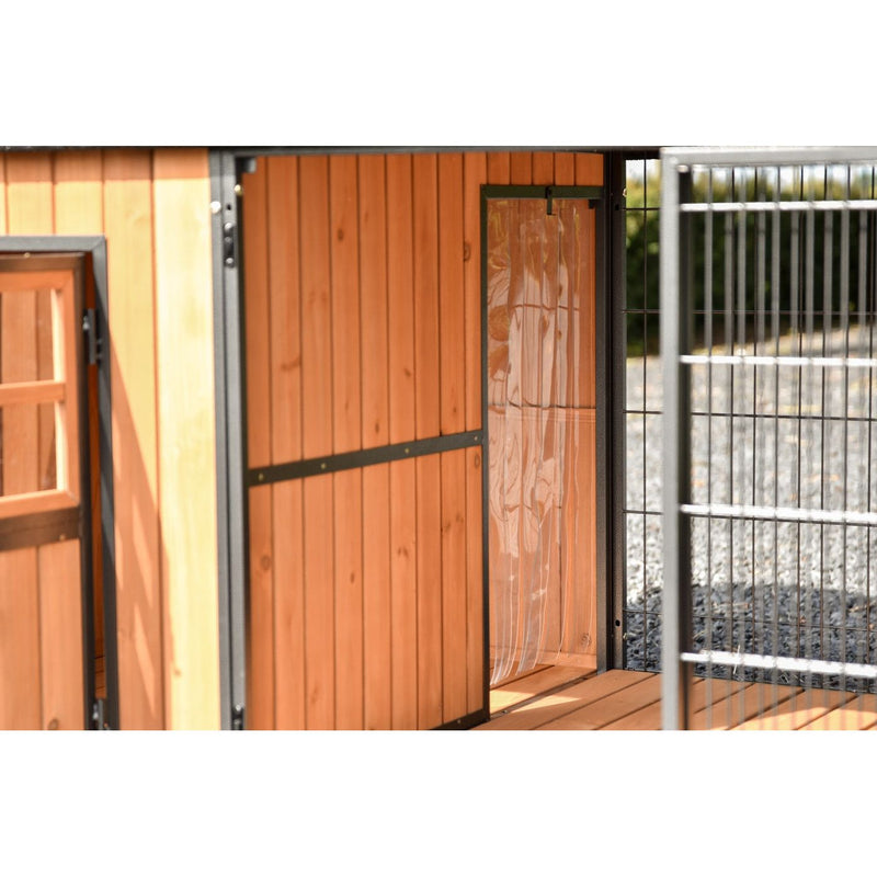 Holzstall aus vertikalen Holzbrettern mit schwarzen Metallbeschlägen, geöffnetem Türblatt und angrenzendem Metallgitterzaun im Aussenbereich.