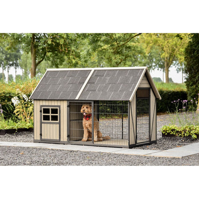 Grosse Holzhundehütte mit Schindeldach und Drahtgitterfront im Garten; ein Hund mit rotem Halsband sitzt im Auslauf hinter dem Gittertor, umliegende Kieswege und Grünflächen.