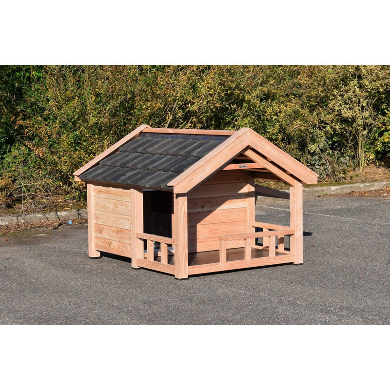 Unbehandeltes Holz-Hundehaus mit offener Veranda, Dach aus schwarzen Dachschindeln, steht auf einer Asphaltfläche vor grünem Buschwerk.