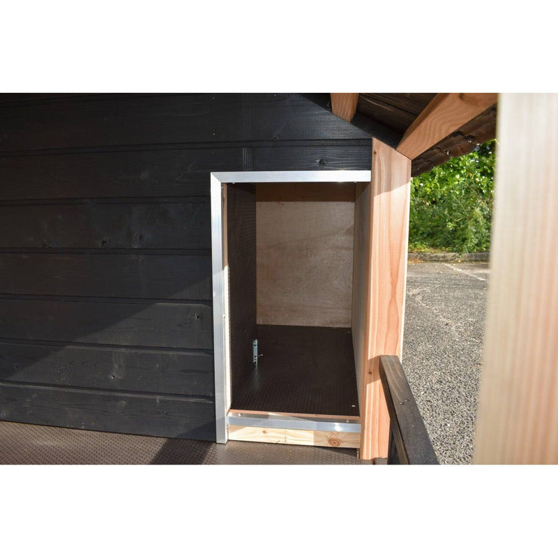 Offener kleiner Outdoor-Speicher oder Schrank aus Holz mit Aluminiumrahmen vor einer schwarzen Holzwand; Innenraum aus Holz, Bodenplatte sichtbar, Gartenzugang im Hintergrund.