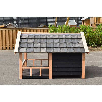 Holz-Hundehütte mit grauem Ziegeldach, rechte dunkle Wand, offener Eingang links, steht auf Asphalt im Garten mit Zaun und grünem Gebüsch im Hintergrund.