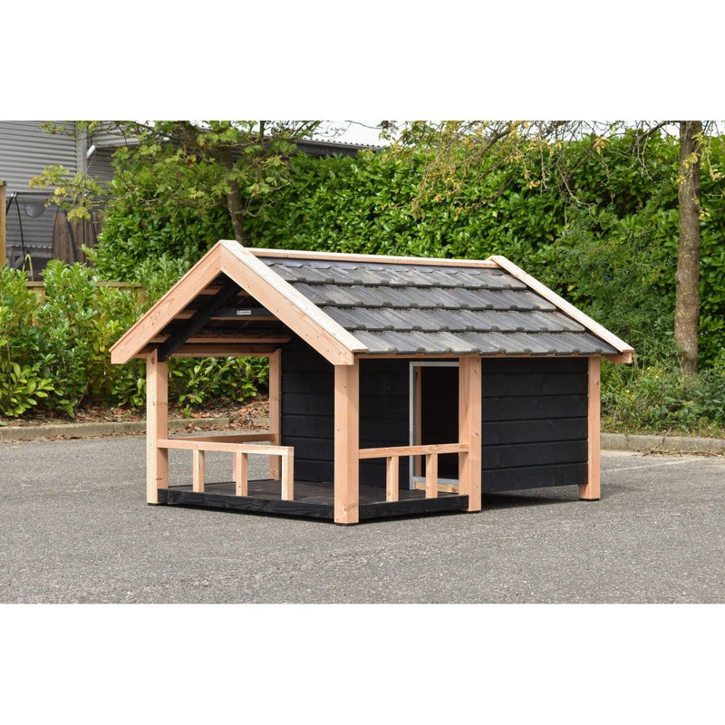 Kleines Garten-Spielhaus aus naturbelassenem Holz mit schwarzen Wänden, Dach aus dunklen Ziegeln, offener Vorderbereich mit Geländer, steht auf Asphalt vor grüner Hecke und Bäumen.