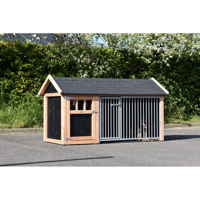 Grosser Outdoor-Hundezwinger aus Holz mit schwarzer Seitenwand und Metallgittertür; Hund sichtbar hinter den Stäben, auf Asphalt vor grüner Hecke.