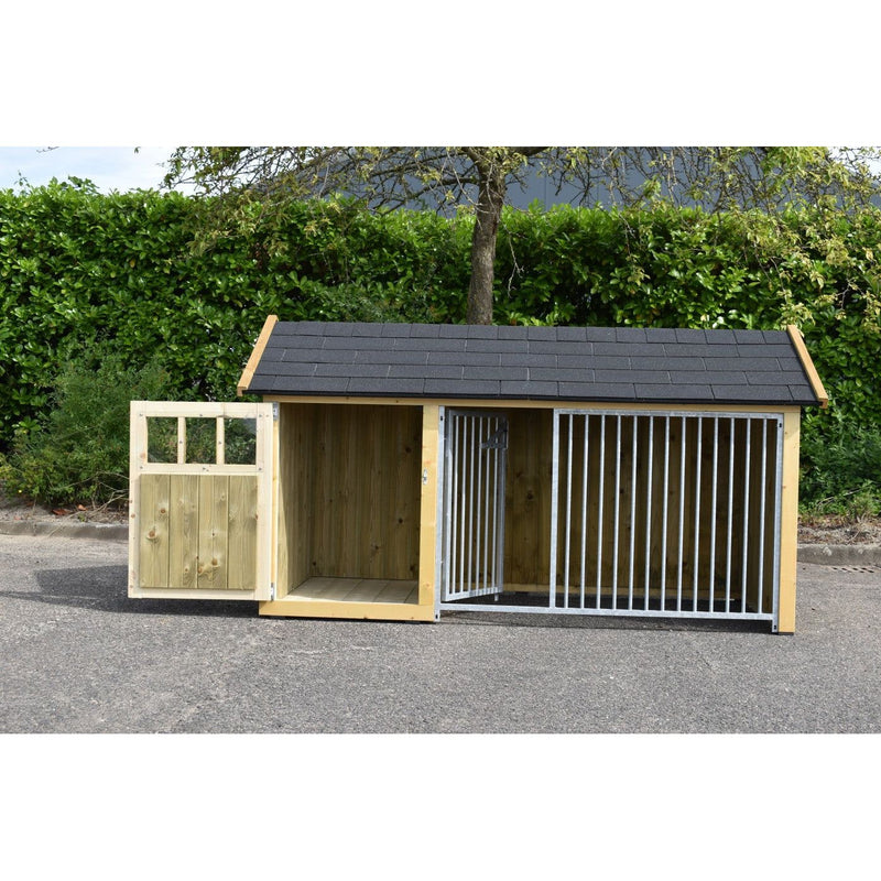 Grosse Hundehütte aus Holz mit zwei Bereichen: links Tür mit Fenster, rechts Zwinger aus vertikalen Metallstäben; dunkles Dach; grüne Hecke und Baum im Hintergrund; Asphaltboden.