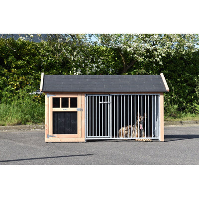Hundezwinger aus Holz mit dunklem Schindeldach; rechter Bereich mit Metallstangen-Gitter, Hund liegt hinter dem Gitter auf asphaltiertem Boden vor einer grünen Hecke.