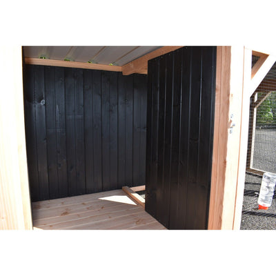 Innenraum eines kleinen Holzschuppens mit schwarzen vertikalen Holzwänden, hellem Holzboden und Türrahmen; aussen Drahtgitter sichtbar.