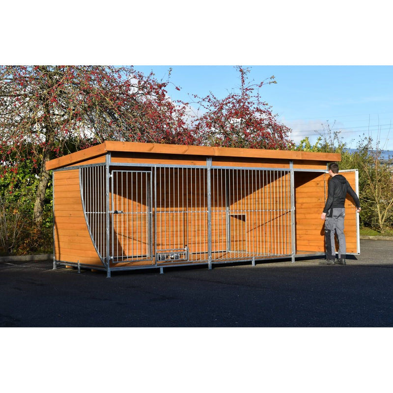 Grosse Hundekäfiganlage aus Holz und Metallrahmen im Freien; eine Person öffnet die Tür rechts; Hintergrund mit Bäumen und blauem Himmel.