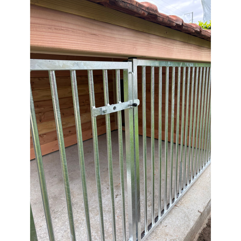 Verzinktes Metallgeländer mit vertikalen Stäben und seitlichem Riegelverschluss; Holzwand im Hintergrund und rotes Ziegeldach darüber.