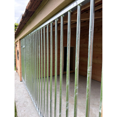 Galvanisierter Stahlzaun mit vertikalen Stäben vor einer Holzhütte – seitliche Gartenaufnahme auf Betonboden.