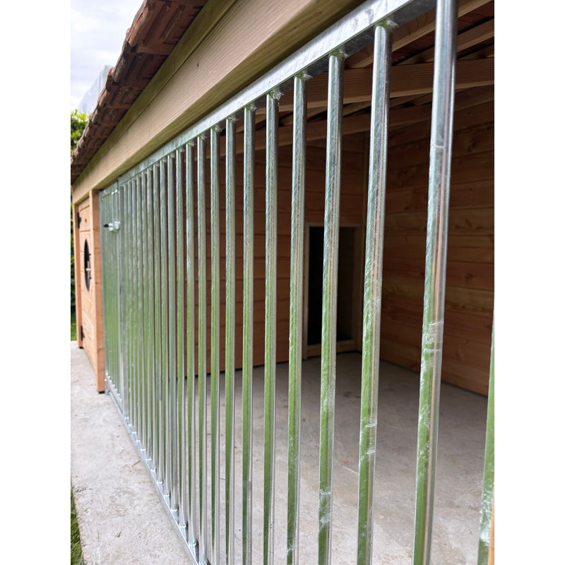 Galvanisierter Stahlzaun mit vertikalen Stäben vor einer Holzhütte – seitliche Gartenaufnahme auf Betonboden.