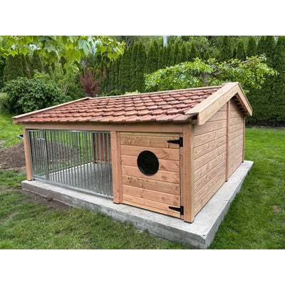 Holz-Hundehütte im Garten mit rotem Ziegeldach und angrenzendem Metallgehege aus Stahlstangen, runder schwarzer Fensterausschnitt in der Tür, auf einer Betonplatte, umgeben von Gras, Bäumen und Hecken.