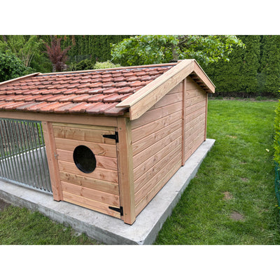 Holz-Hundehütte mit rotem Tonziegeldach auf einer Betonplatte im Garten; naturbelassene Holzwände, runde schwarze Eingangsluke in der Tür, grüne Rasenfläche und Hecken im Hintergrund.