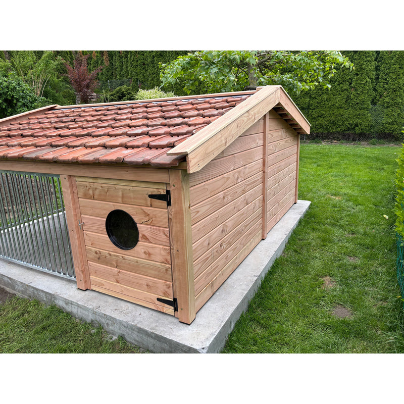 Holz-Hundehütte mit rotem Tonziegeldach auf einer Betonplatte im Garten; naturbelassene Holzwände, runde schwarze Eingangsluke in der Tür, grüne Rasenfläche und Hecken im Hintergrund.