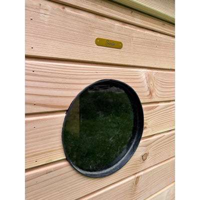 Rundes schwarzes Fenster (Portloch) in heller Holzwand mit horizontalen Brettern, daneben ein goldfarbenes Messingschild mit handgeschriebenem Schriftzug.