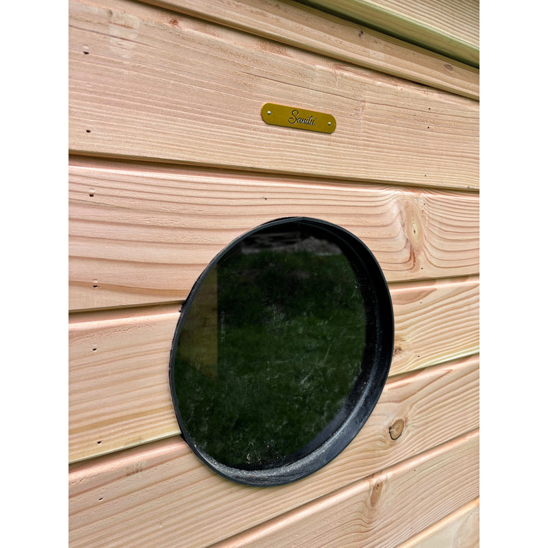 Rundes schwarzes Fenster (Portloch) in heller Holzwand mit horizontalen Brettern, daneben ein goldfarbenes Messingschild mit handgeschriebenem Schriftzug.