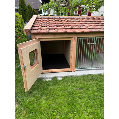 Holz-Hundehütte mit rotem Ziegeldach im Garten; offene linke Tür mit rundem Fenster, dunkler Innenraum, rechts Metall-Gittertor, steht auf einer Betonplatte neben Gras.