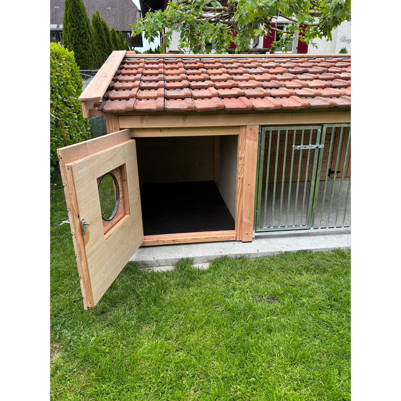 Holz-Hundehütte mit rotem Ziegeldach im Garten; offene linke Tür mit rundem Fenster, dunkler Innenraum, rechts Metall-Gittertor, steht auf einer Betonplatte neben Gras.