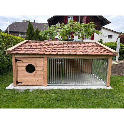 Holz-Hundekäfig mit rotem Ziegeldach und Metallgitterfront, linke Tür mit rundem Fenster, steht auf einer Betonplatte im Garten vor Wohnhäusern.