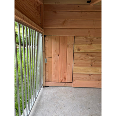 Kleines Holztürchen in einer Holzwand eines Holzhauses; Tür aus senkrechten Brettern mit Metallriegel, links Metallgeländer und Gras im Aussenbereich, Betonboden im Vordergrund.