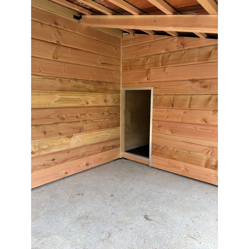 Innenansicht eines Holzschuppens mit horizontalen Holzwänden, sichtbaren Dachbalken und einer kleinen rechteckigen Öffnung in der Wand, die zu einem dunklen kleinen Raum führt; Betonboden.