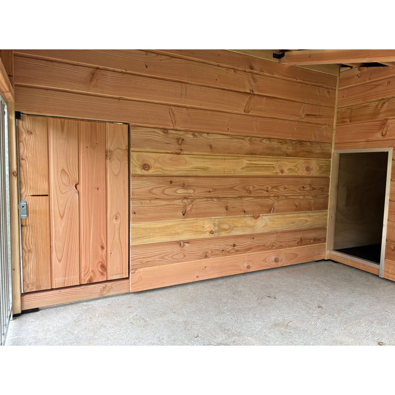 Innenraum eines holzverkleideten Gartenhauses mit horizontalen Holzbrettern, links eine geschlossene Holztür, rechts ein offener Durchgang, Betonboden.