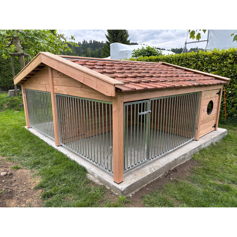 Grosser Hundezwinger aus Holz mit vertikalen Metallstäben, rotem Ziegel-Dach, seitliches rundes Lochfenster, steht auf Betonfundament im Garten.