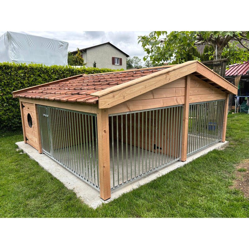 Grosser Outdoor-Hundezwinger aus Holz mit Metallstäben und rotem Ziegeldach, steht auf einer Betonplatte im Garten.