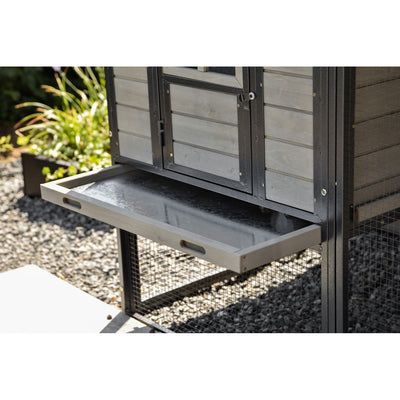 Hühnerstall mit ausziehbarer Metall-Schublade im Frontbereich, schwarzer Rahmen, Holzverkleidung, Drahtgitter und Kiesboden im Garten.