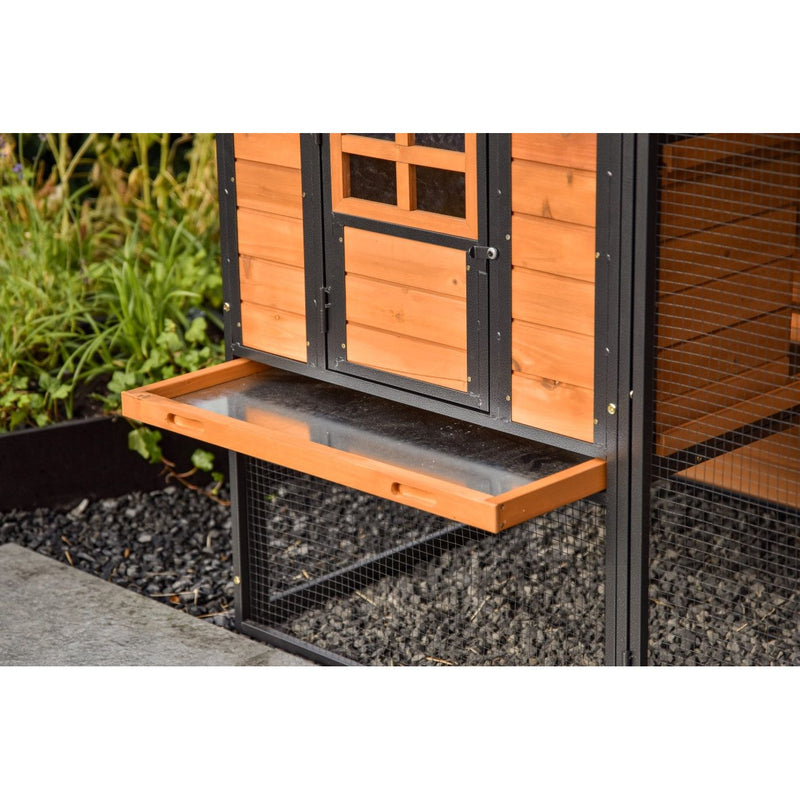 Hühnerstall aus Holz mit schwarzem Metallrahmen, herausziehbare Ablage vor der Tür, Drahtgitterboden, Garten im Hintergrund.