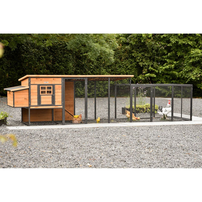 Hühnerstall aus Holz mit Drahtgehege und länglicher Laufzone; mehrere Hühner laufen im Gehege auf Kiesboden vor grünem Hintergrund.