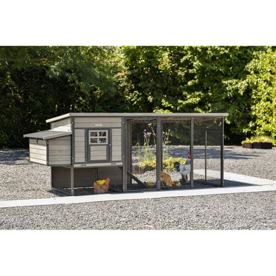 Holz-Hühnerstall mit Drahtgehege und freiem Auslauf auf Kiesboden, umgeben von Bäumen; mehrere Hühner und ein Hahn im Gehege, linker Anbau des Stalls.