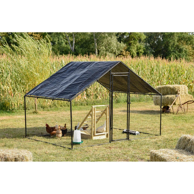 Hühnerstall-Gehege aus schwarzem Metallrahmen mit Planendach, auf einer Wiese neben Heuballen; Hühner im Gehege, sichtbare Futter- und Wasserbehälter, Maisfeld und Bäume im Hintergrund