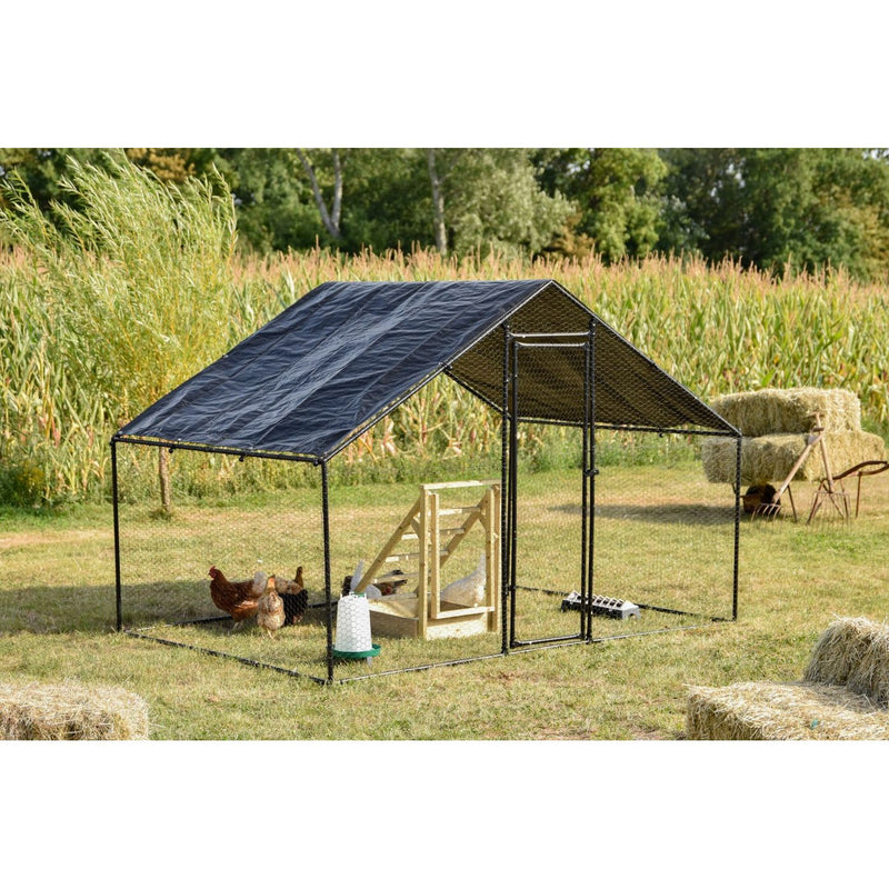 Hühnerstall-Gehege aus schwarzem Metallrahmen mit Planendach, auf einer Wiese neben Heuballen; Hühner im Gehege, sichtbare Futter- und Wasserbehälter, Maisfeld und Bäume im Hintergrund